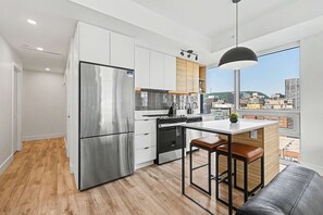 A modern kitchen with a stainless steel refrigerator, wooden cabinets, a bar stool, and a city view through large windows. - 2 Bedroom/2 Bathroom Suite (Montreal)