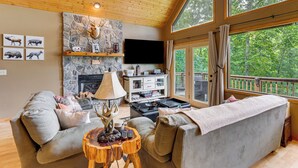 Ein gemütliches Wohnzimmer mit Kamin aus Naturstein, einem Flachbildfernseher und einem großen Fenster mit Blick auf einen Wald.