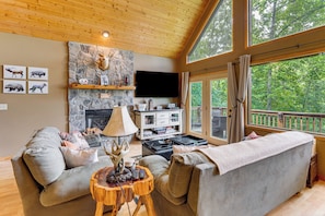 Ein gemütliches Wohnzimmer mit Kamin aus Naturstein, einem Flachbildfernseher und einem großen Fenster mit Blick auf einen Wald.