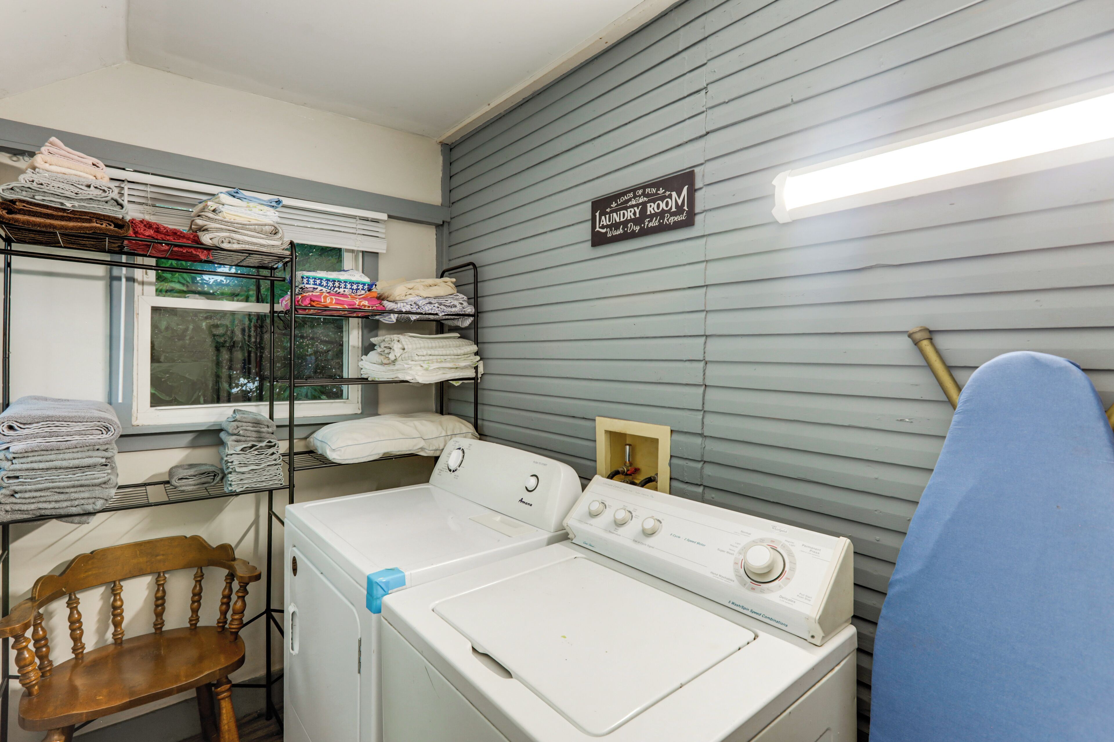 A laundry room with a front-loading washer and dryer, shelves with folded towels, and a sign indicating laundry services.