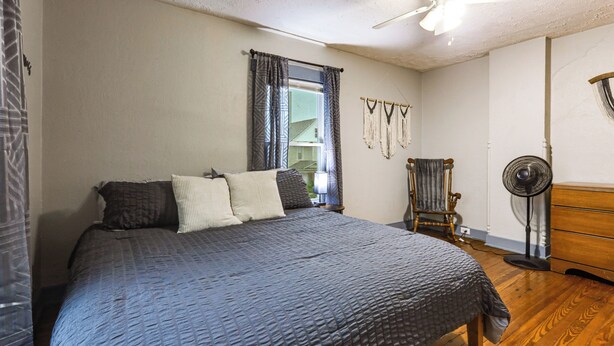 A bedroom with a large bed, a rocking chair, a standing fan, and a window with curtains.