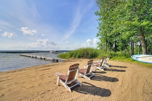 A beach with several wooden Adirondack chairs, a dock extending into calm water, and a row of trees in the background.