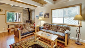 A spacious living room with a high ceiling, a stone fireplace, a large window with blinds, and a patterned sofa set. - Minutes to slopes & shore! Cozy cabin w/ hot tub (South Lake Tahoe)