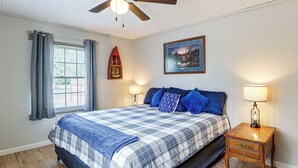 A bedroom with a bed, a nightstand, a lamp, a window with curtains, a ceiling fan, and a wall-mounted shelf with decorative items. - Walk to Park & Creek! Spearfish Couple’s Retreat (Spearfish)