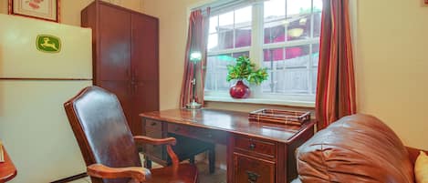 Una habitación con escritorio de madera, una silla de cuero, un refrigerador, un armario, una ventana con cortinas y una planta.