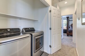 A laundry room with two front-loading washers and dryers, a white door leading to another room, and a hallway with a carpeted floor. - Relax Poolside, Explore South Mtn! Extended Escape (Phoenix)