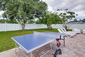 Ein Tischtennis-Tisch steht auf einer Ziegelterrasse, im Hintergrund ist ein weißer Zaun und Bäume zu sehen.