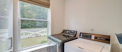 A laundry room with a washing machine and dryer, a window with a view of a garden, and a door with a handle.
