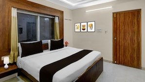 Quarto de hotel com cama, cabeceira de madeira, janela com cortinas, mesa de cabeceira com abajur e porta.