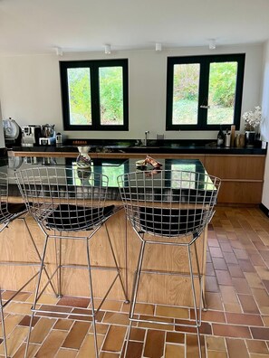 A modern kitchen with a central island, two wireframe chairs, and a view of greenery through the windows. - Villa Hardelot, near the golf course and the beach (Neufchâtel-Hardelot)