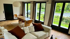 En moderne stue med sofa, spisebord, stole og store vinduer med udsigt over et grønt landskab.