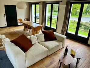 A modern living room with a sofa, dining table, chairs, and large windows offering a view of a green landscape. - Villa Hardelot, near the golf course and the beach (Neufchâtel-Hardelot)