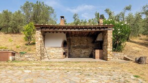 A stone-built outdoor structure with a wooden roof and two arched openings, surrounded by dry grass and olive trees. - Casale le Lupe Apartments - Ground Floor (Campiglia Marittima)