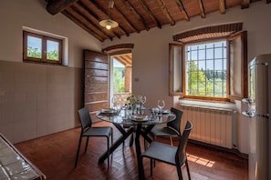 A dining area with a round table, four chairs, a window with a view of greenery, a radiator, and a ceiling with exposed beams. - Casale le Lupe Apartments – First Floor (Campiglia Marittima)