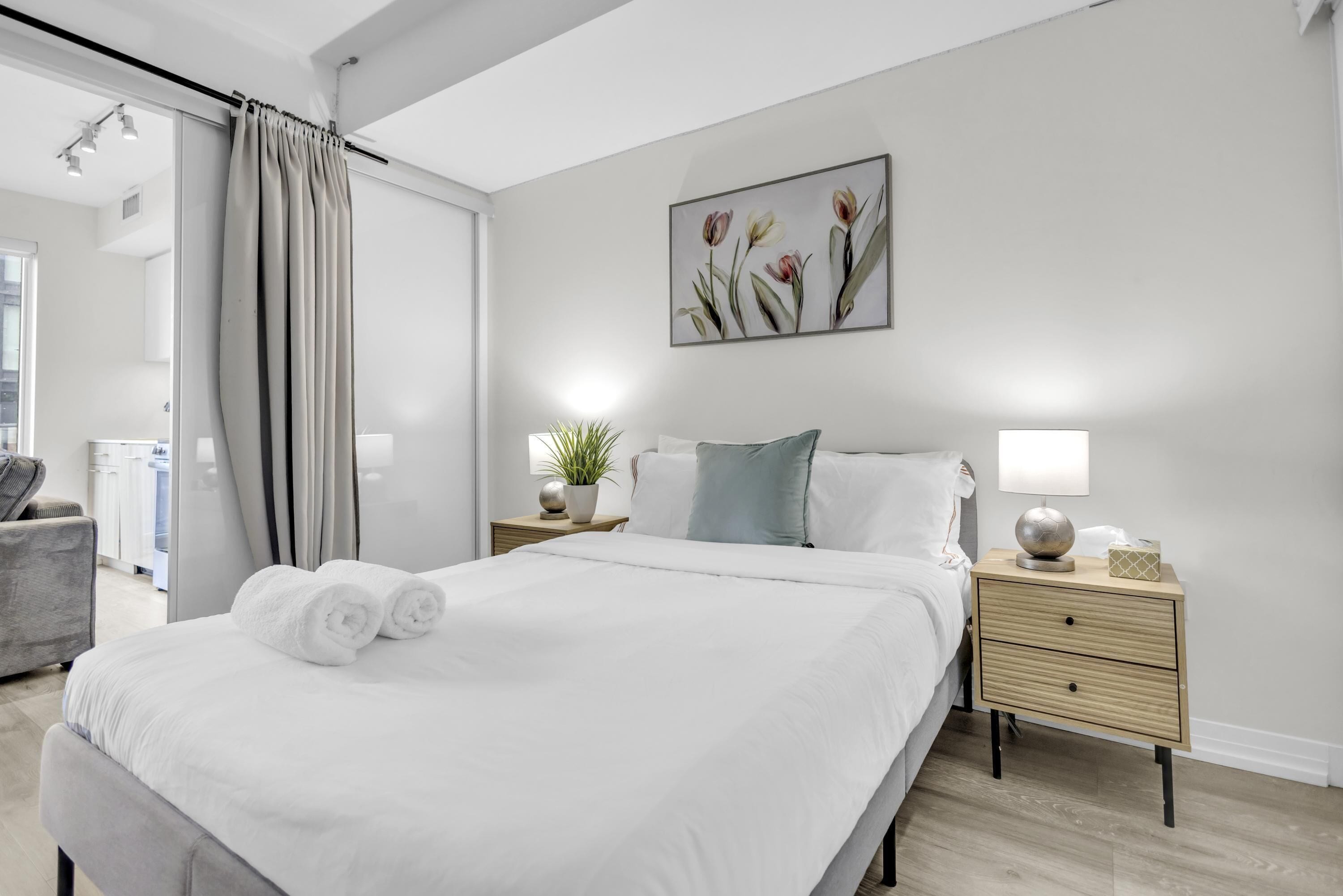 A modern bedroom with a large bed, a nightstand with a lamp, a potted plant, and a painting on the wall.