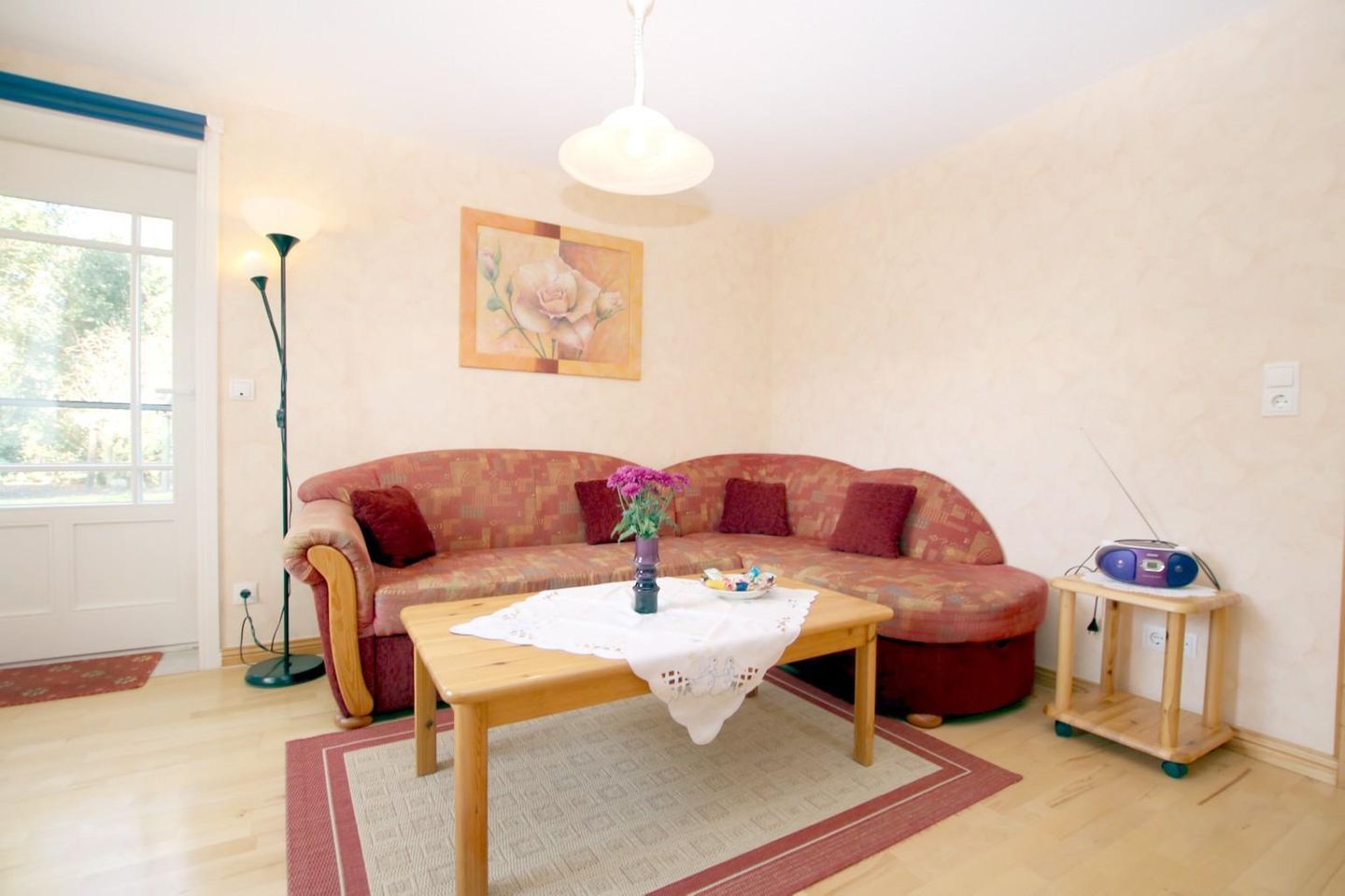 Uma sala de estar com um sofá, uma mesa de centro de madeira, um abajur, um quadro e um rádio em uma mesinha lateral.