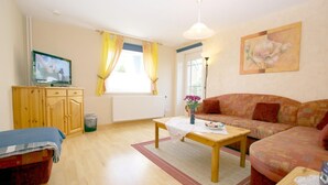 A furnished living room with a sofa, a television, a wooden cabinet, and a window with curtains. - Babendörpstieg 20, Apt. 2 - 3-room vacation apartment in Nieblum, approx. 75 square meters, on the ground floor, with (Nieblum)