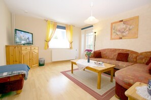 Ein möbliertes Wohnzimmer mit Sofa, Fernseher, Holzschrank und einem Fenster mit Vorhängen.