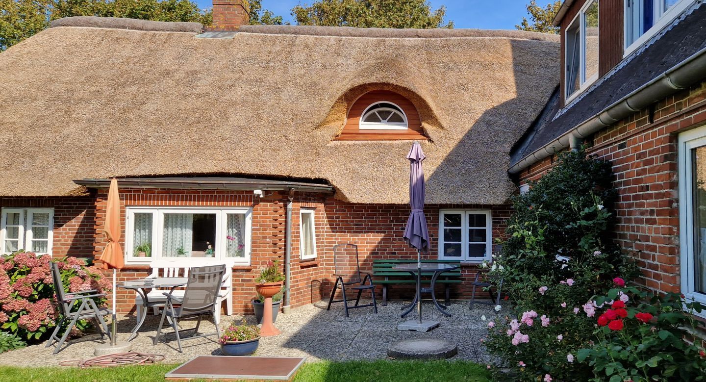 Een rieten huis met een bakstenen gevel, een terras met tuinmeubilair, en een bloemrijke tuin.