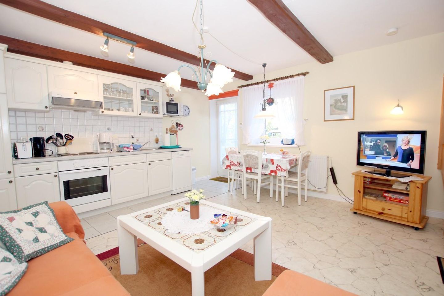 Een moderne keuken met witte kasten, een eethoek met tafel en stoelen, een televisie op een houten standaard, en een gezellige woonkamer met een bank en salontafel.