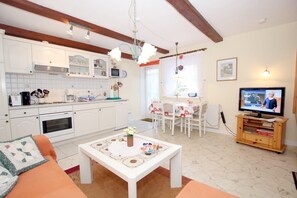 Eine moderne Küche mit weißen Schränken, ein Essbereich mit Tisch und Stühlen, ein Fernseher auf einem Holzgestell und ein gemütliches Wohnzimmer mit Sofa und Couchtisch.