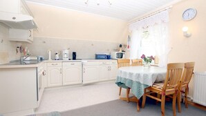 Een kleine keuken met witte kastjes, een eettafel met stoelen, een klok aan de muur en een raam met gordijnen.