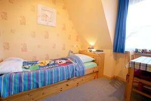 A single bed with a colorful Mickey Mouse blanket, a bedside table with a lamp, and a window with curtains. - Babendörpstieg 20, Apt. 3 - 4-room vacation apartment in Nieblum, approx. 65 square meters, on the 1st floor (Nieblum)