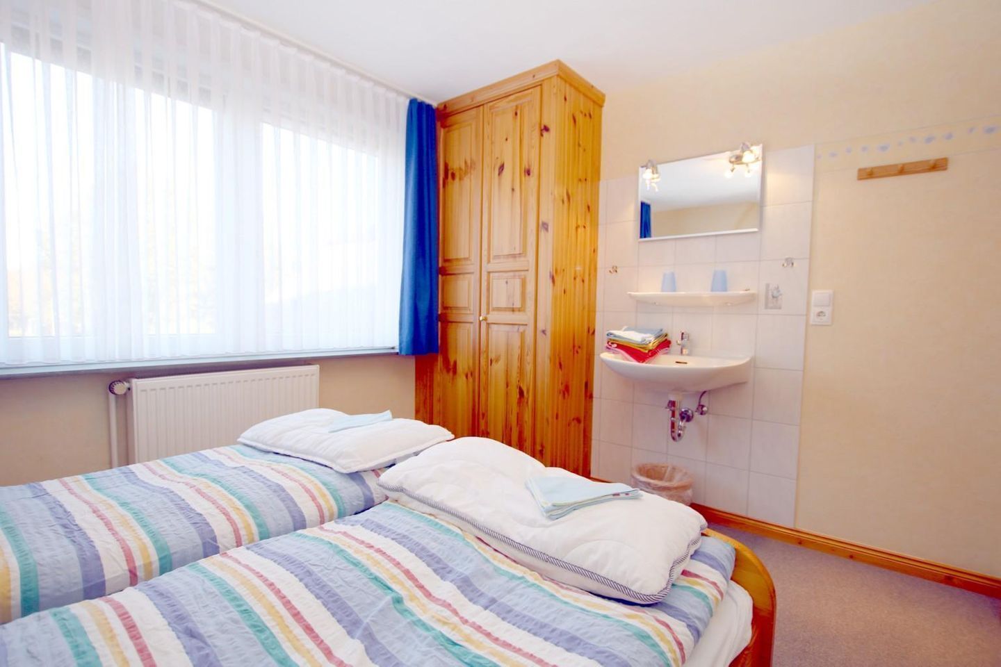 A bedroom with a bed, a wooden wardrobe, a window with curtains, and a bathroom with a sink and mirror.