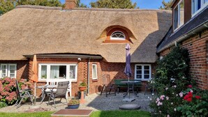 Een rieten dak huis met een bakstenen gevel, een terras met tuinmeubilair, en een bloemen tuin.