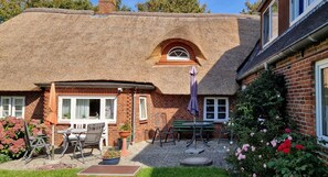 A thatched-roof house with a brick exterior, a patio with outdoor furniture, and a garden with flowers. - Babendörpstieg 20, Apt. 3 - 4-room vacation apartment in Nieblum, approx. 65 square meters, on the 1st floor (Nieblum)