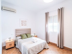 Ein Schlafzimmer mit einem Bett, Nachttischen, einer Wand-Klimaanlage, einem Fenster mit Vorhängen und einem Wandbild.