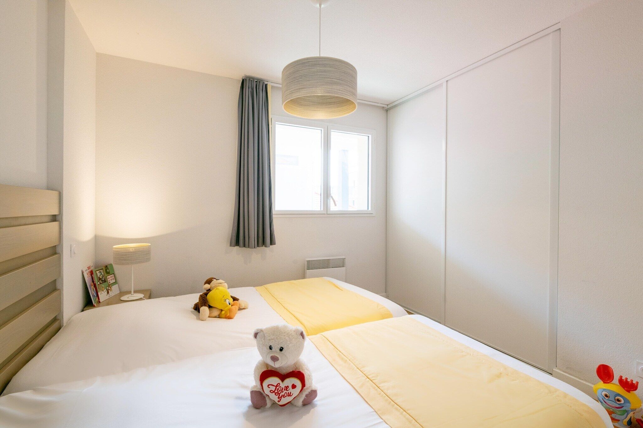 Quarto de hotel com cama, abajur, janela com cortinas e um urso de pelúcia na cama.
