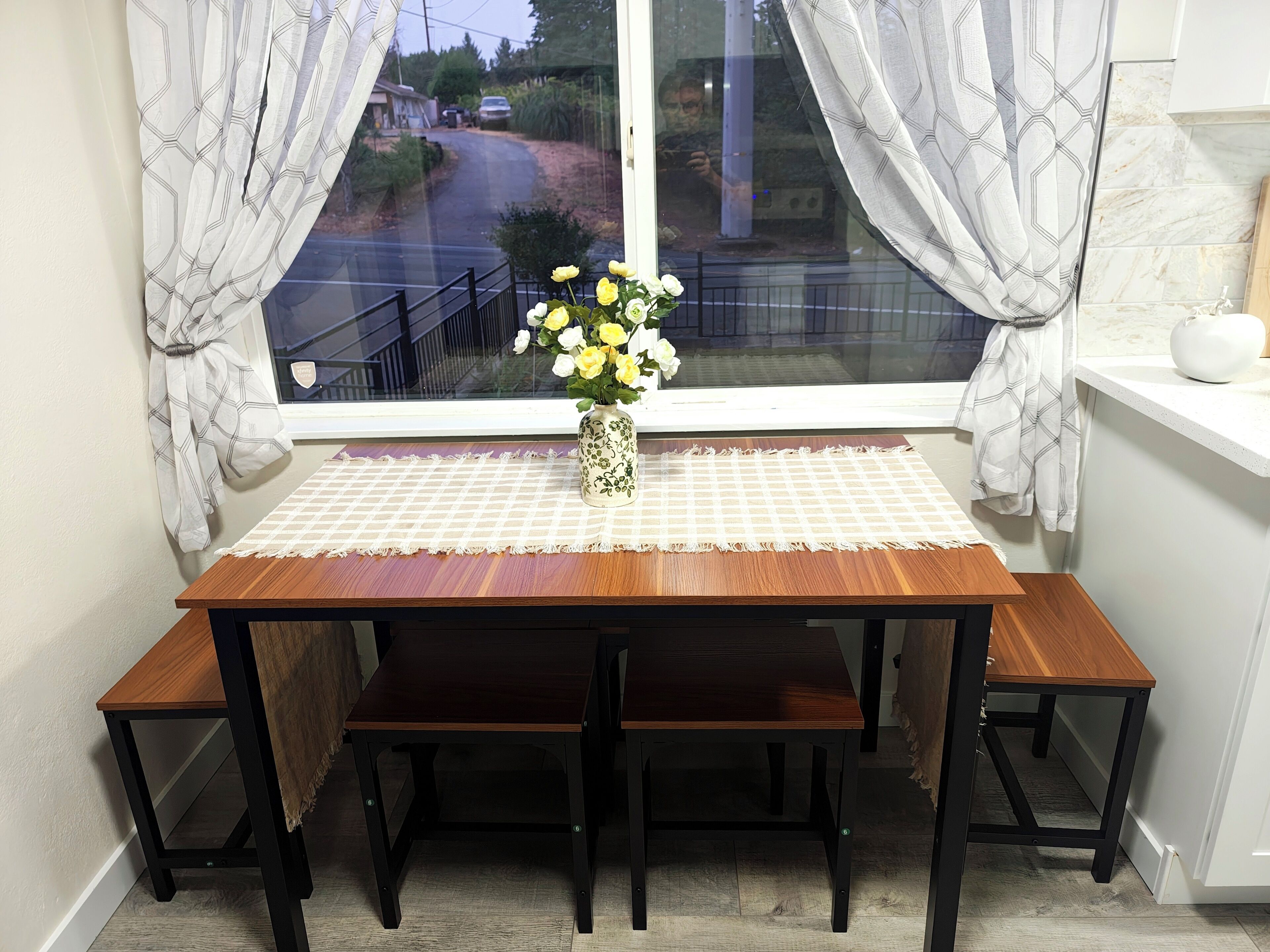 A wooden dining table with benches, a vase with flowers, and a view of a road through a window with curtains.