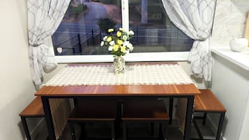 A wooden dining table with benches, a vase with flowers, and a view of a road through a window with curtains.