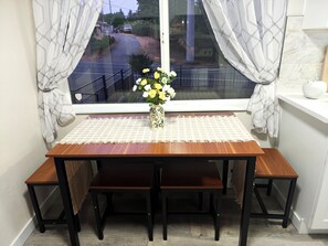 A wooden dining table with benches, a vase with flowers, and a view of a road through a window with curtains.