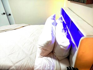 A bed with white bedding and a blue headboard, a bedside table, and a wall-mounted shelf.