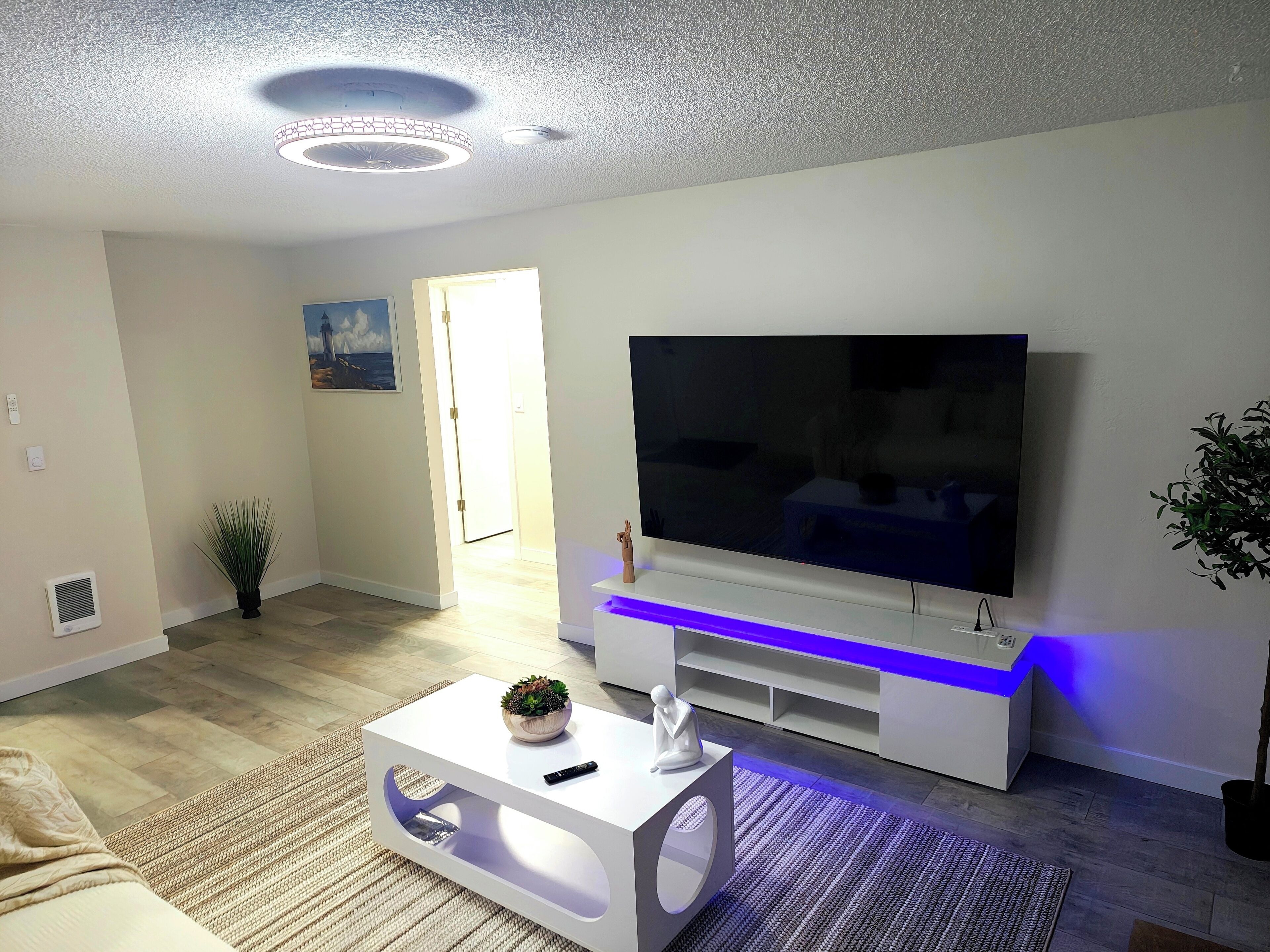 A modern living room with a flat-screen TV, a white entertainment unit with blue LED lighting, a white coffee table with a plant, and a large window with natural light.