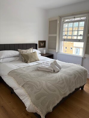 A neatly made bed with a quilt, pillows, and folded towels. There is a bedside table with a lamp and a framed picture. A window with shutters is visible, and the room has wooden flooring.