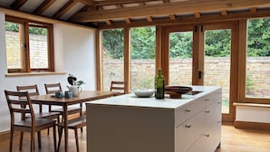 A modern kitchen with a central island, wooden beams, large windows, and a brick exterior wall.