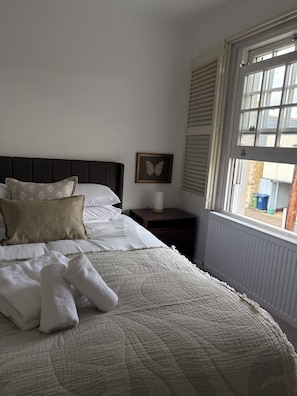 A neatly made bed with a quilt, pillows, and folded towels. There is a bedside table with a lamp and a framed picture on the wall.