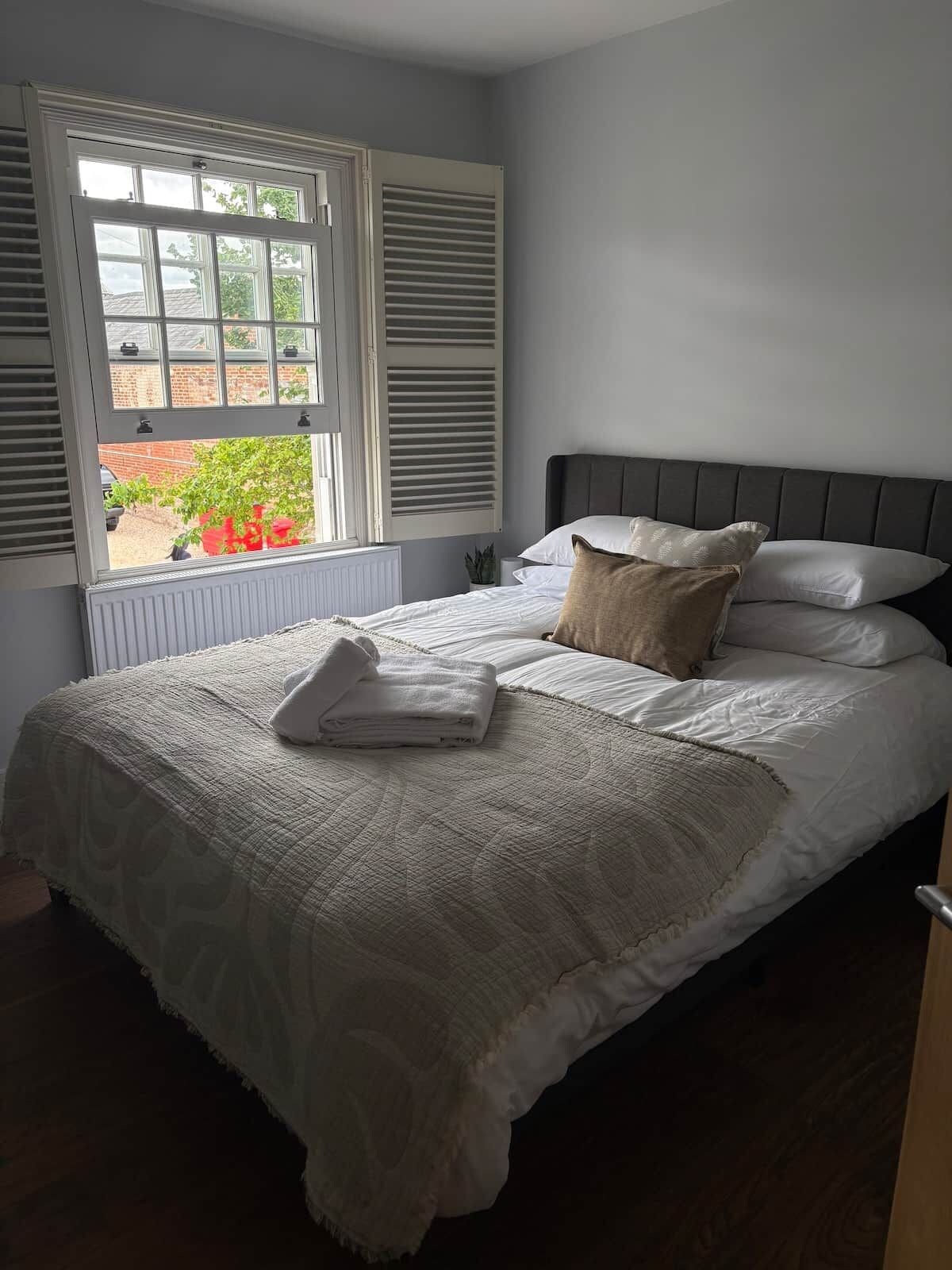 A bedroom with a bed, pillows, a window with shutters, and a view of greenery outside.