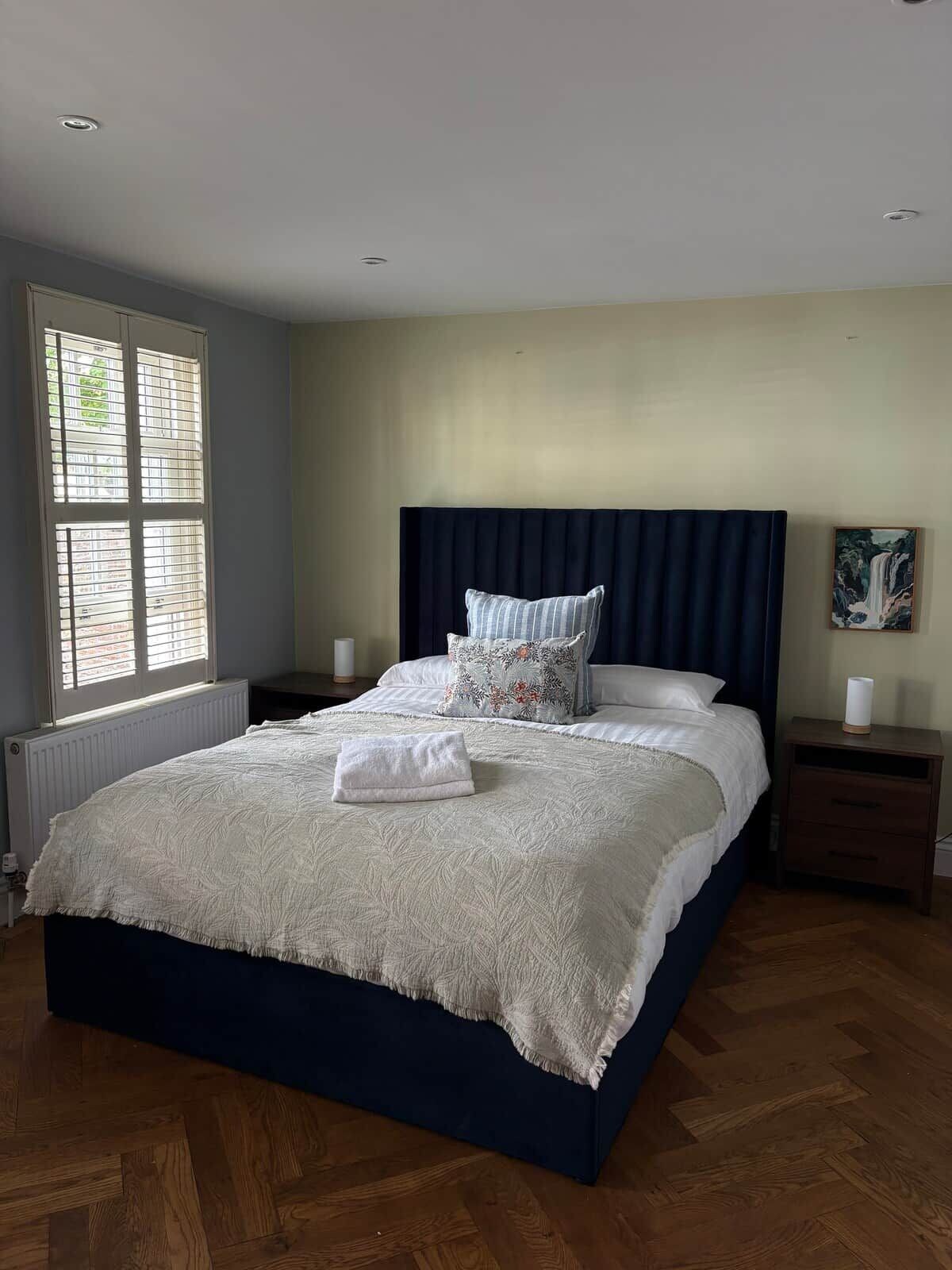 A bedroom with a large bed, a headboard, a nightstand, a window with blinds, and a painting on the wall.