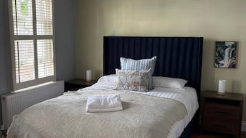A bedroom with a large bed, a headboard, a nightstand, a window with blinds, and a painting on the wall.