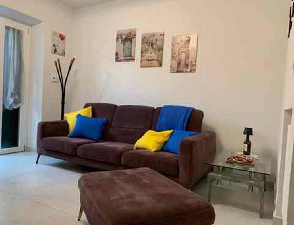 Una sala de estar con un sofá marrón, cojines azules y amarillos, una mesa de centro de vidrio y una puerta que da a un balcón.