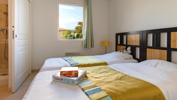Habitación de hotel con dos camas, una ventana con cortinas, cabecera de madera y mesita de noche con lámpara.