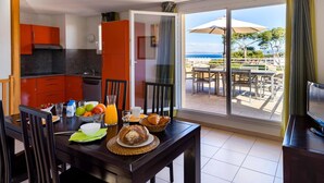 Un comedor con una mesa lista para el desayuno, una cocina con armarios naranjas, y un balcón con mesa y sillas con vistas al mar.