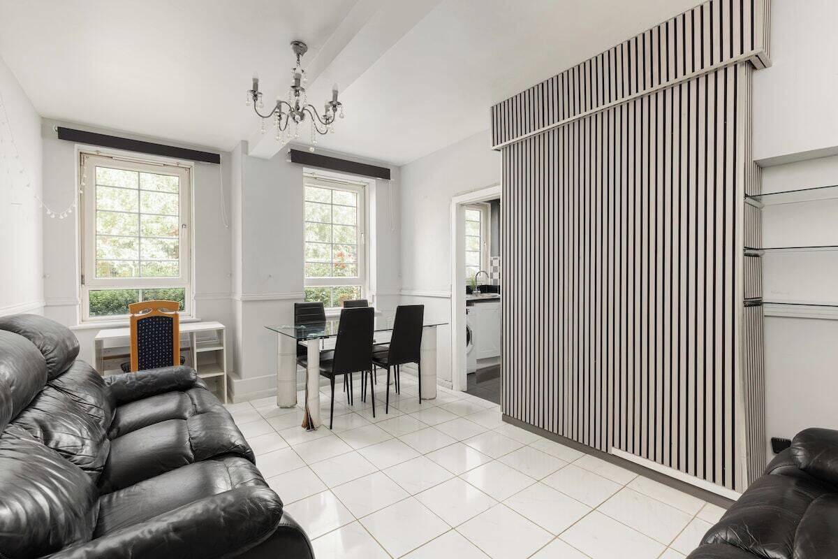A modern living room with a black leather sofa, a dining area with a glass table and chairs, and a kitchen visible through a sliding door.