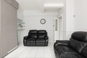 A living room with two black leather sofas, a white tiled floor, a wall clock, and a door leading to another room. - New! Lovely 3BR w/Garden | 6 Mins to Canada Water (Greater London)