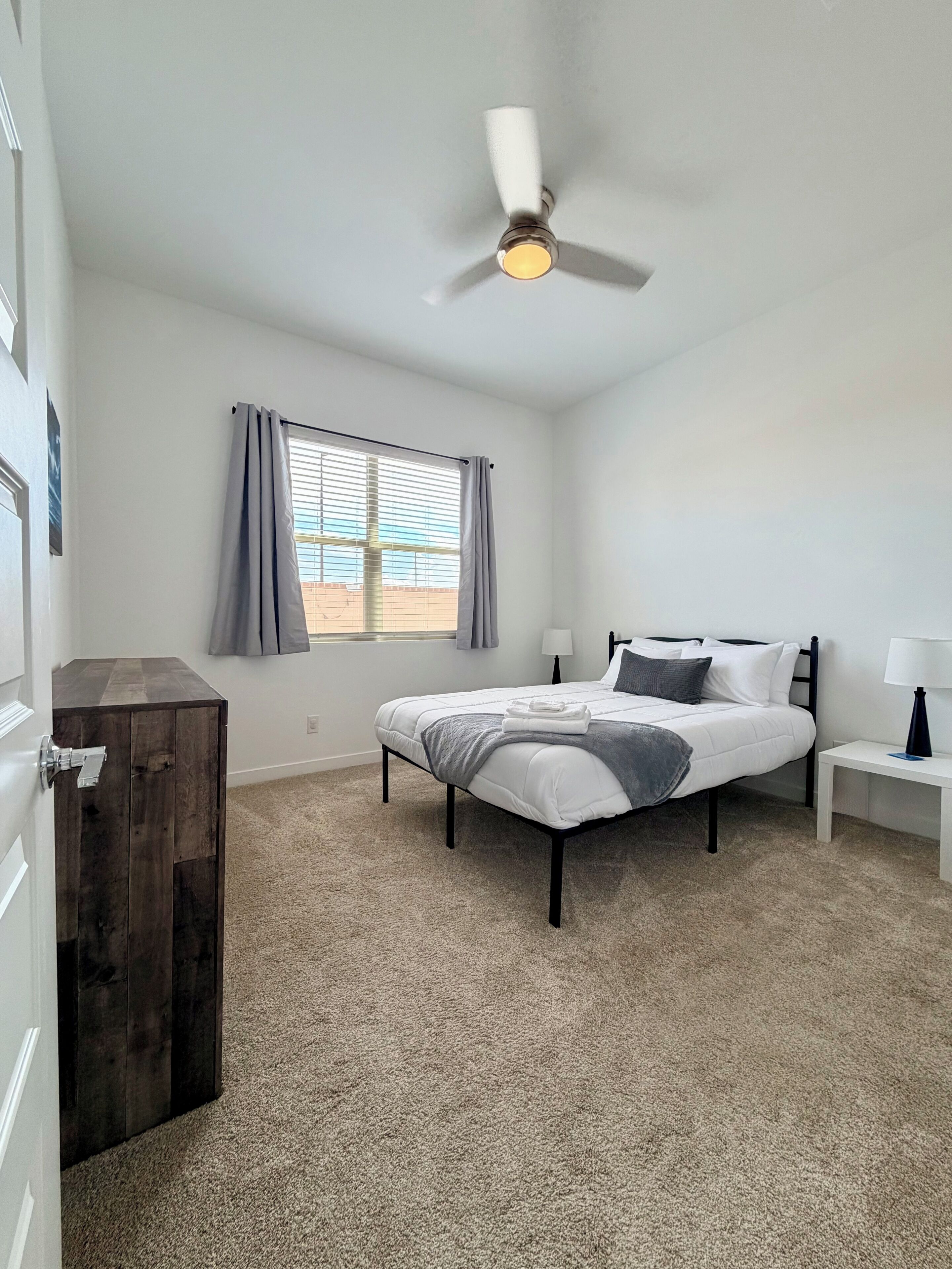 A bedroom with a bed, bedside tables, a ceiling fan, a window with curtains, and a door.
