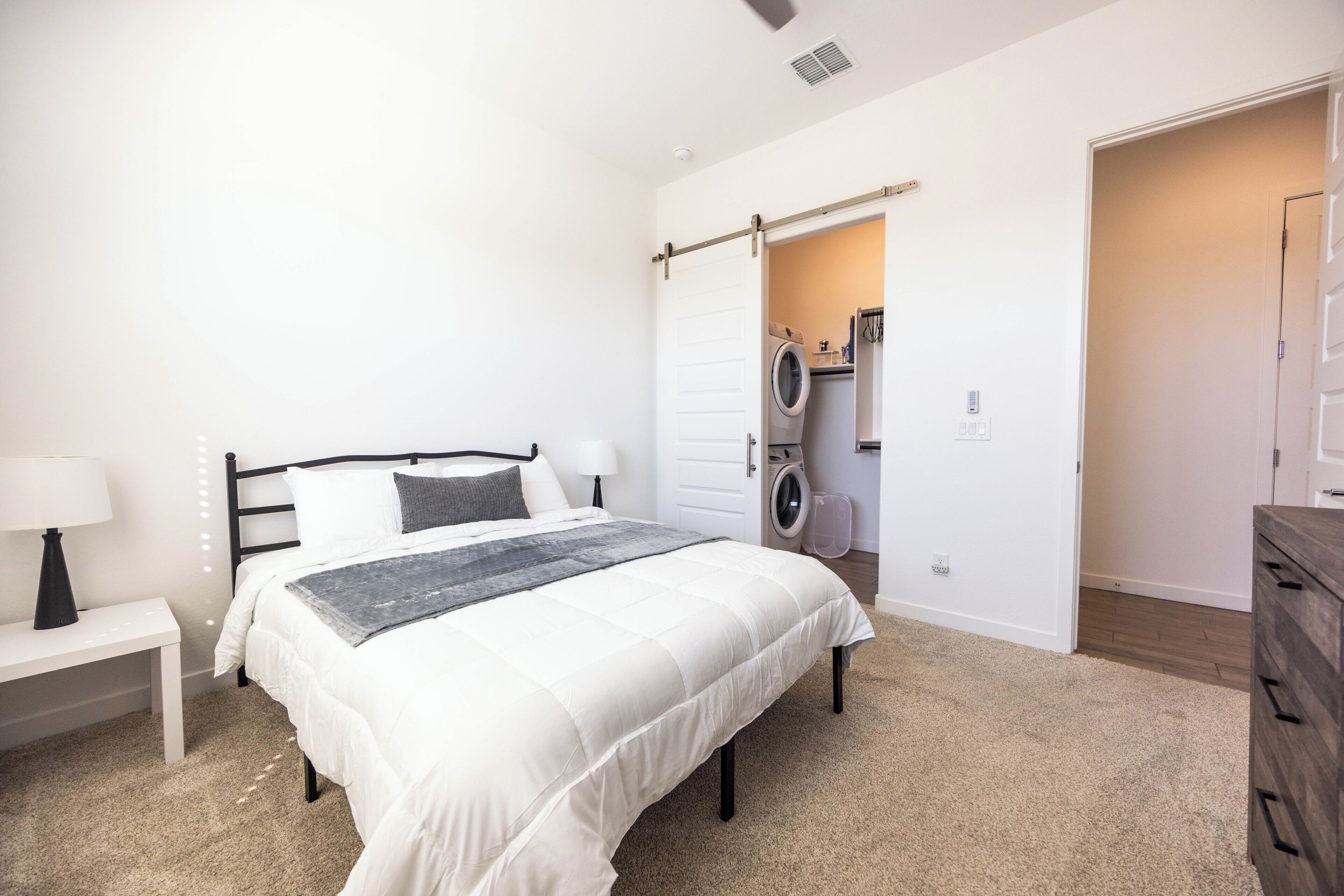 A bedroom with a bed, bedside tables, a washing machine, and a door leading to another room.